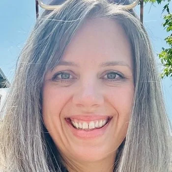Close-up of a woman with long gray hair smiling outdoors with trees and a blue sky in the background.