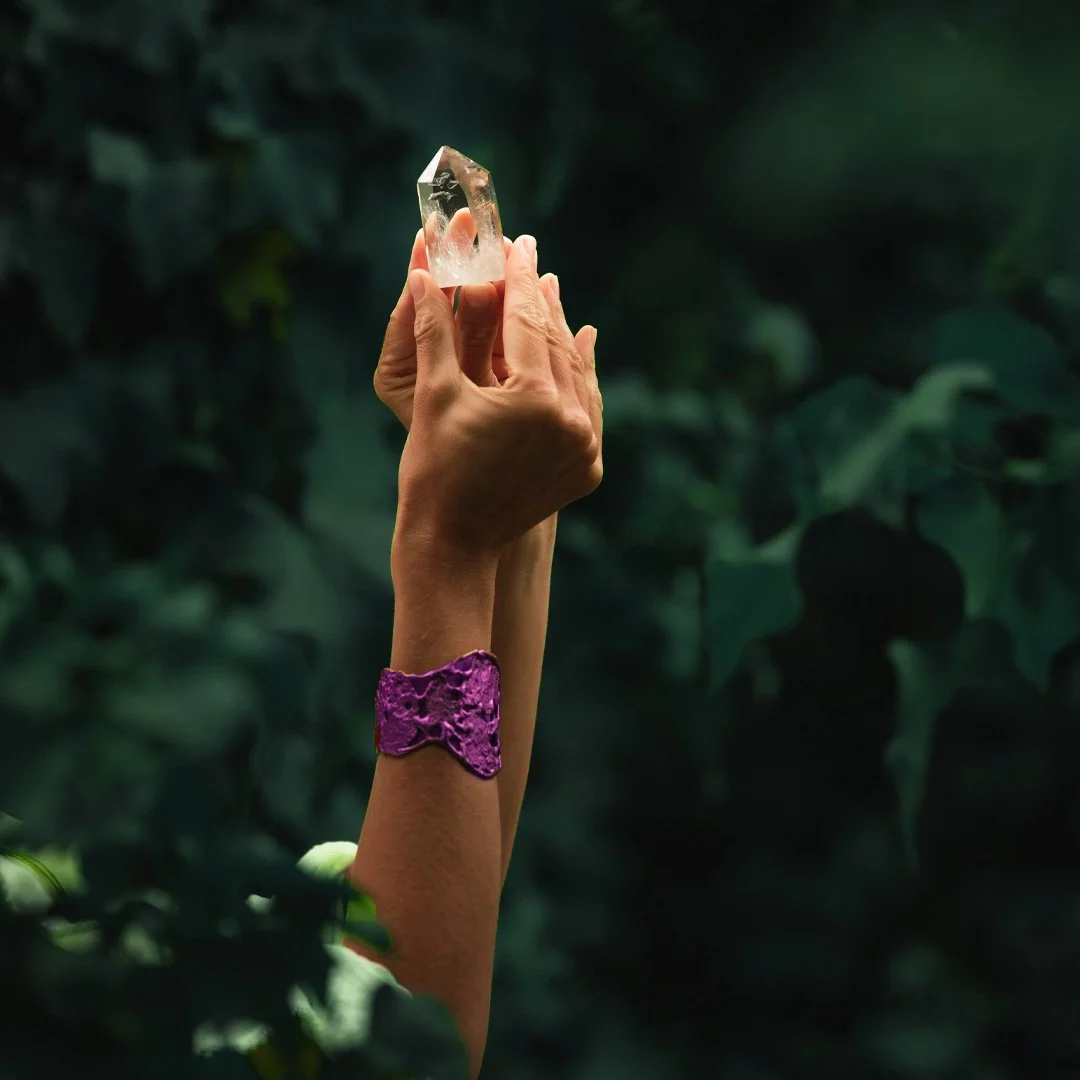Mano sosteniendo un cristal de cuarzo en una mano, con fondo de hojas verdes y una pulsera morada en la muñeca.