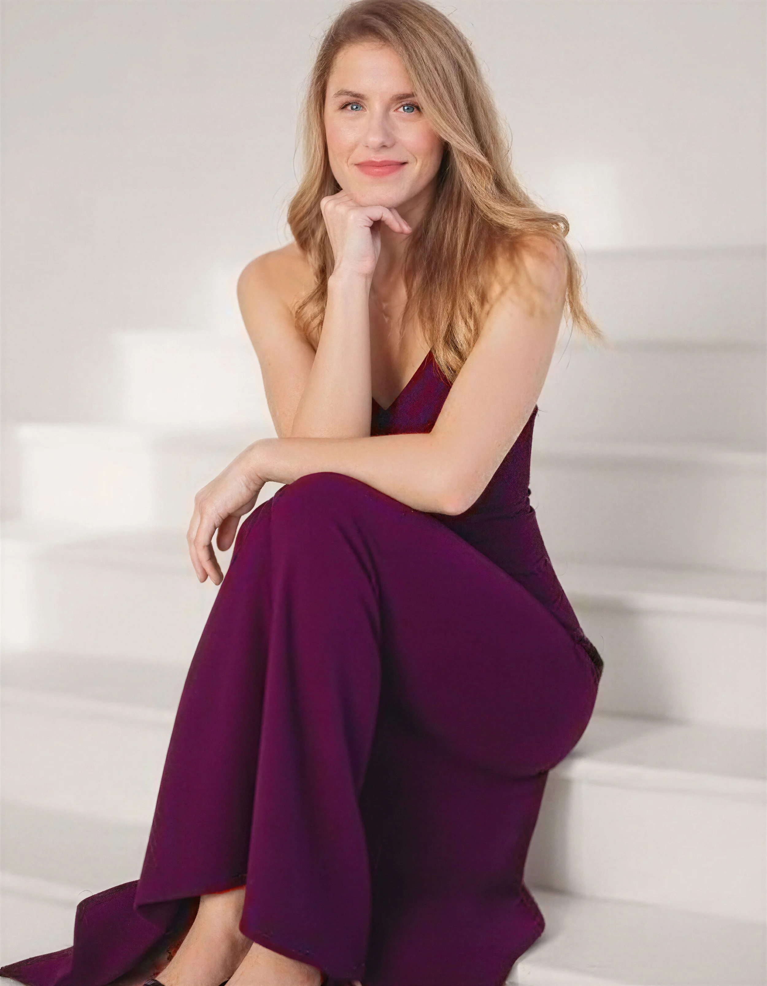 Mujer de cabello rubio y camiseta con pantalón morado, sonriendo mientras se sienta en unas escaleras blancas.