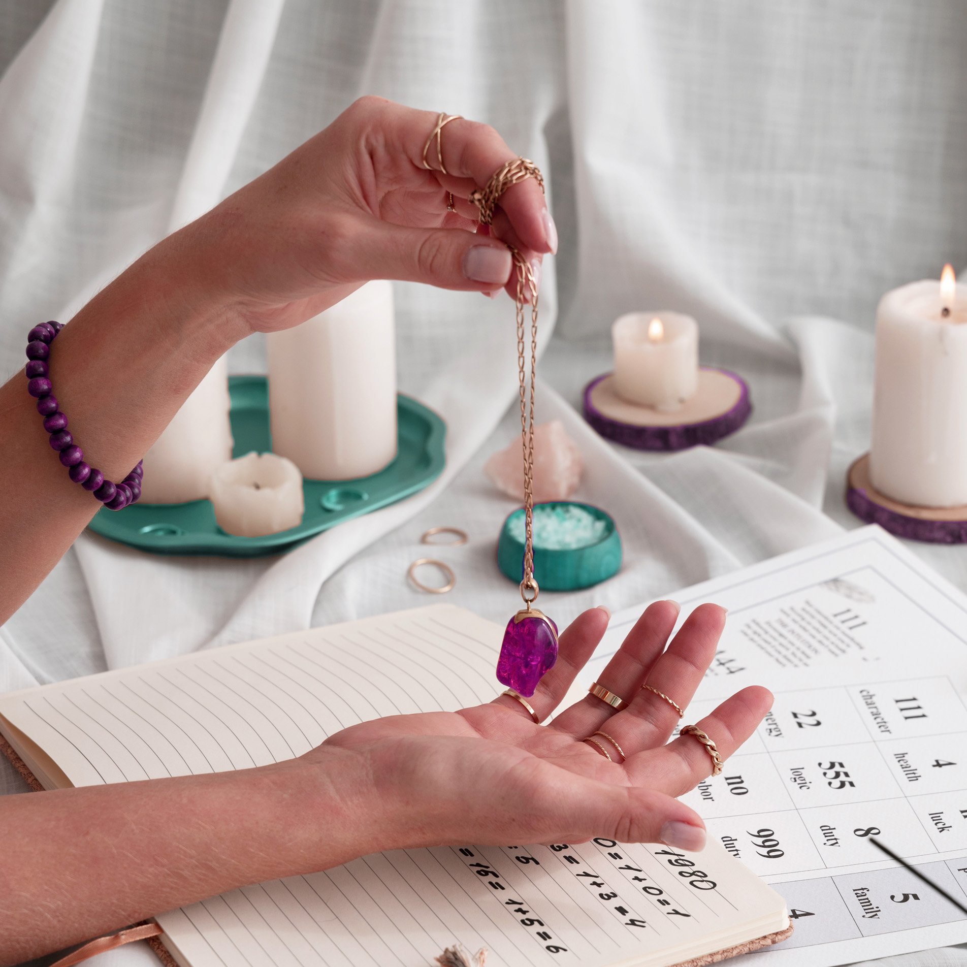 Mano colocando un collar de cadena con una piedra rosada sobre la palma de la mano, en un escritorio con velas encendidas, un cuaderno con notas y un calendario, y varias alianzas doradas.