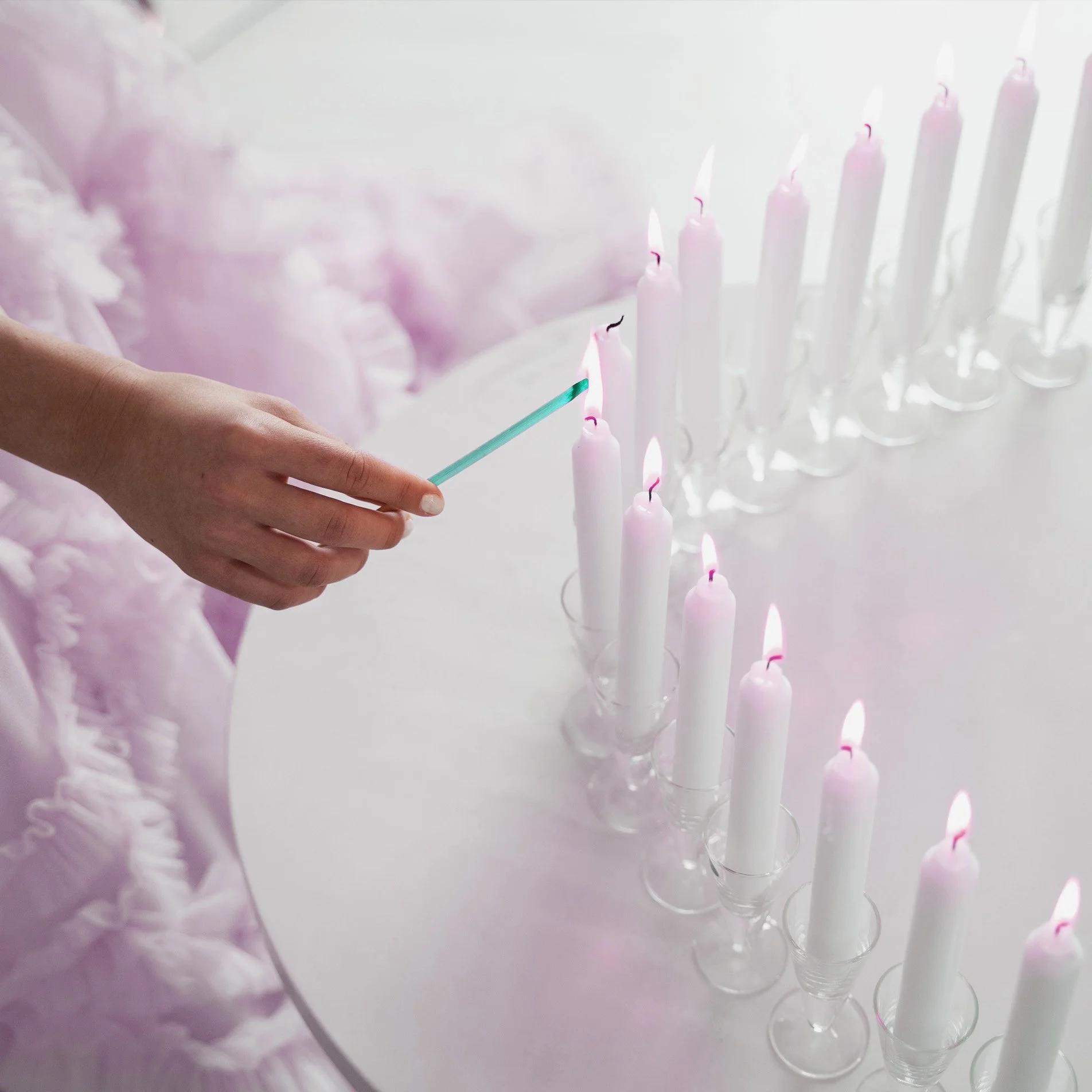 Una mano encende velas blancas en un candelabro sobre una mesa blanca con decoraciones en tonos rosados.
