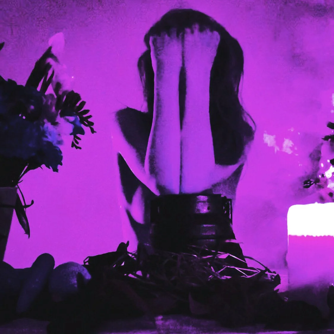 Una mujer con cabello oscuro cubriéndose el rostro con las manos, iluminada con luces moradas y púrpuras, rodeada de plantas y objetos decorativos.