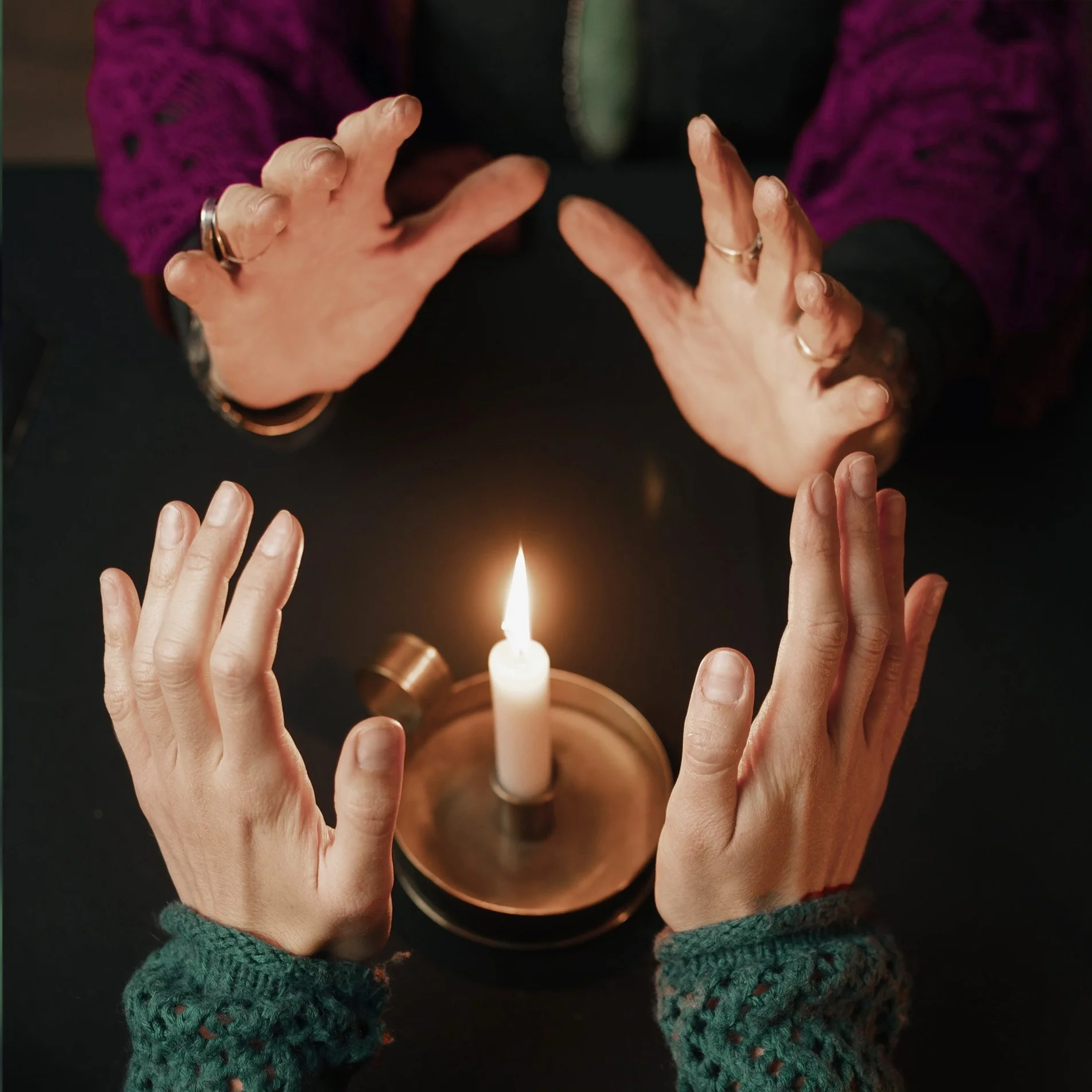 Dos manos adultas con uñas largas y sin esmalte, una con puño cerrado y otra con dedos extendidos, rodeando una vela encendida en un platito de cobre, y una tercera mano, con uñas cortas y sin esmalte, con los dedos extendidos y con calentadores de t