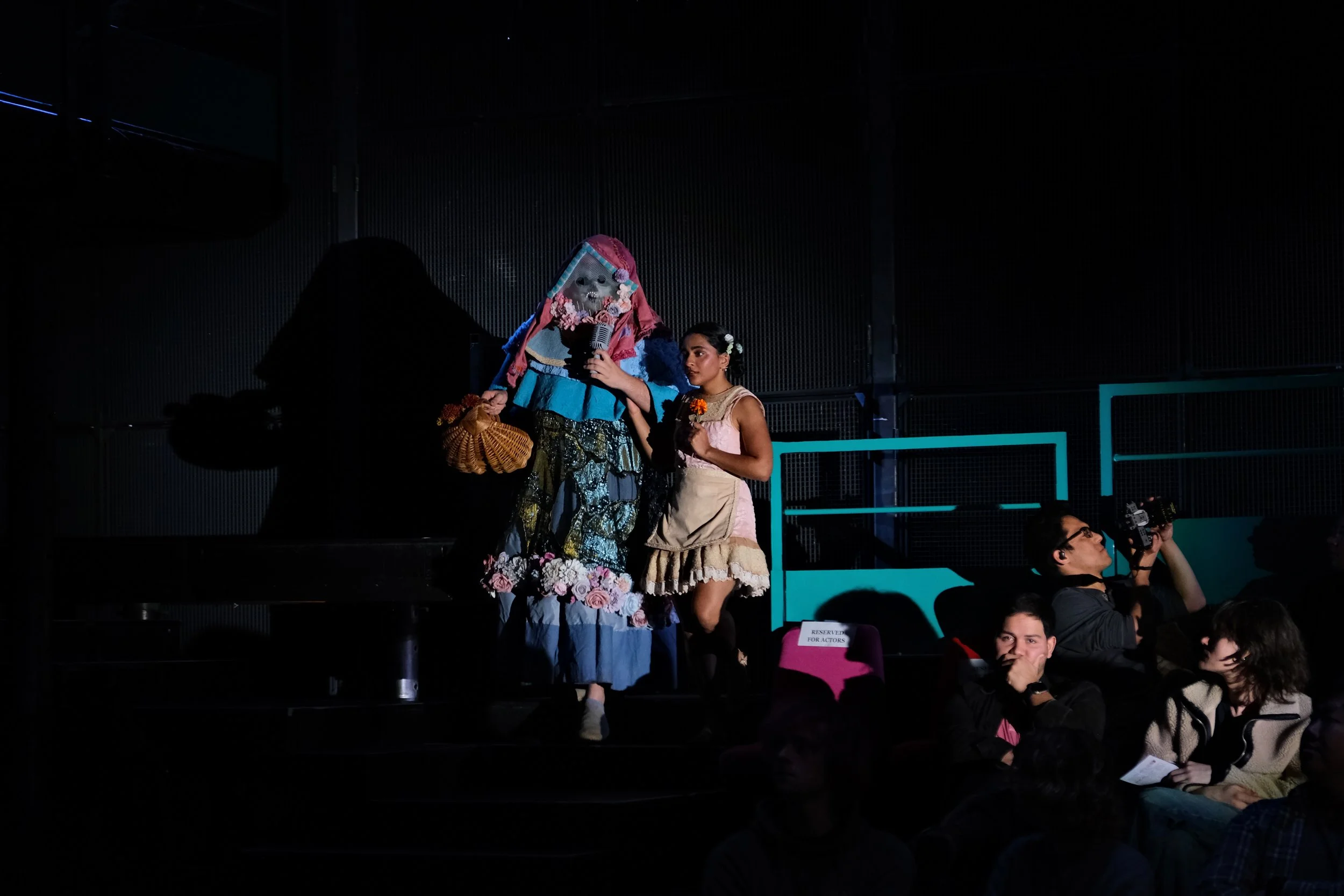 A stage performance featuring a person in a colorful, elaborate costume with a mask, and a young girl holding a flower, with an audience watching.
