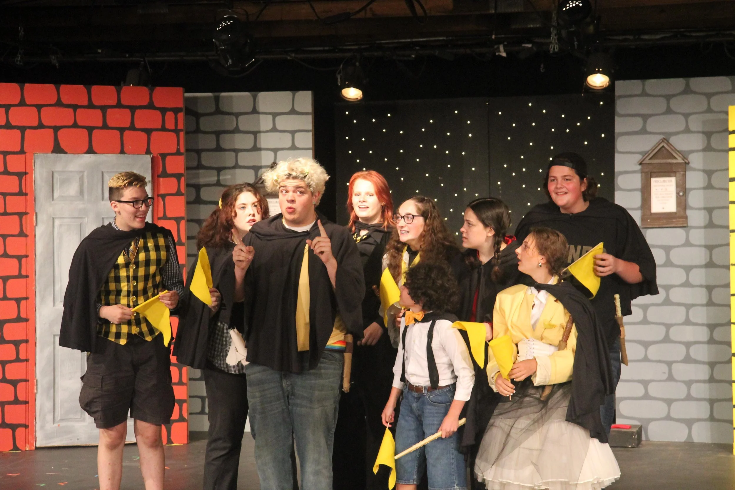 Group of young actors performing on stage in costumes, holding yellow flags, with a backdrop of brick wall and a door, under stage lights.