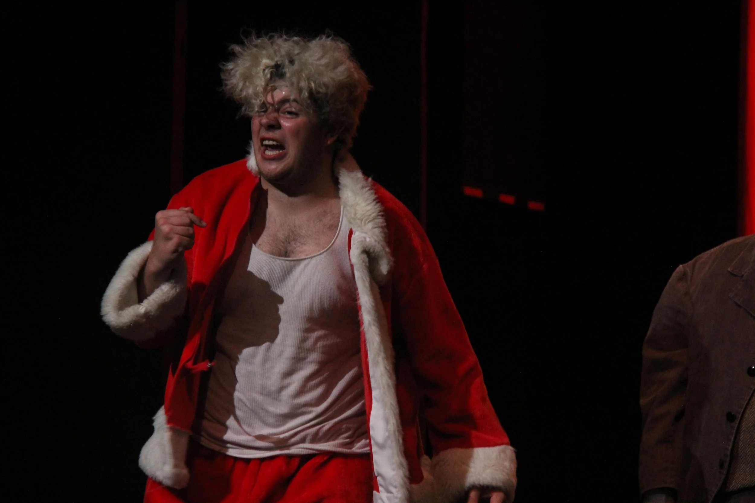 A man with wild, curly blonde hair is wearing a Santa suit and a wet undershirt, and appears to be shouting or yelling on stage.