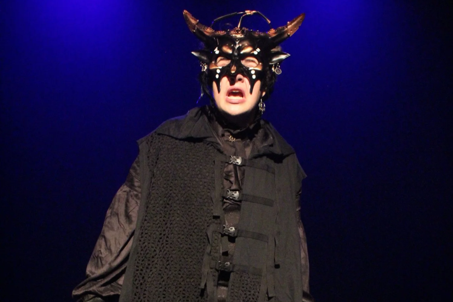 Person in black outfit wearing a black butterfly mask with horns and antennae on stage against dark background.
