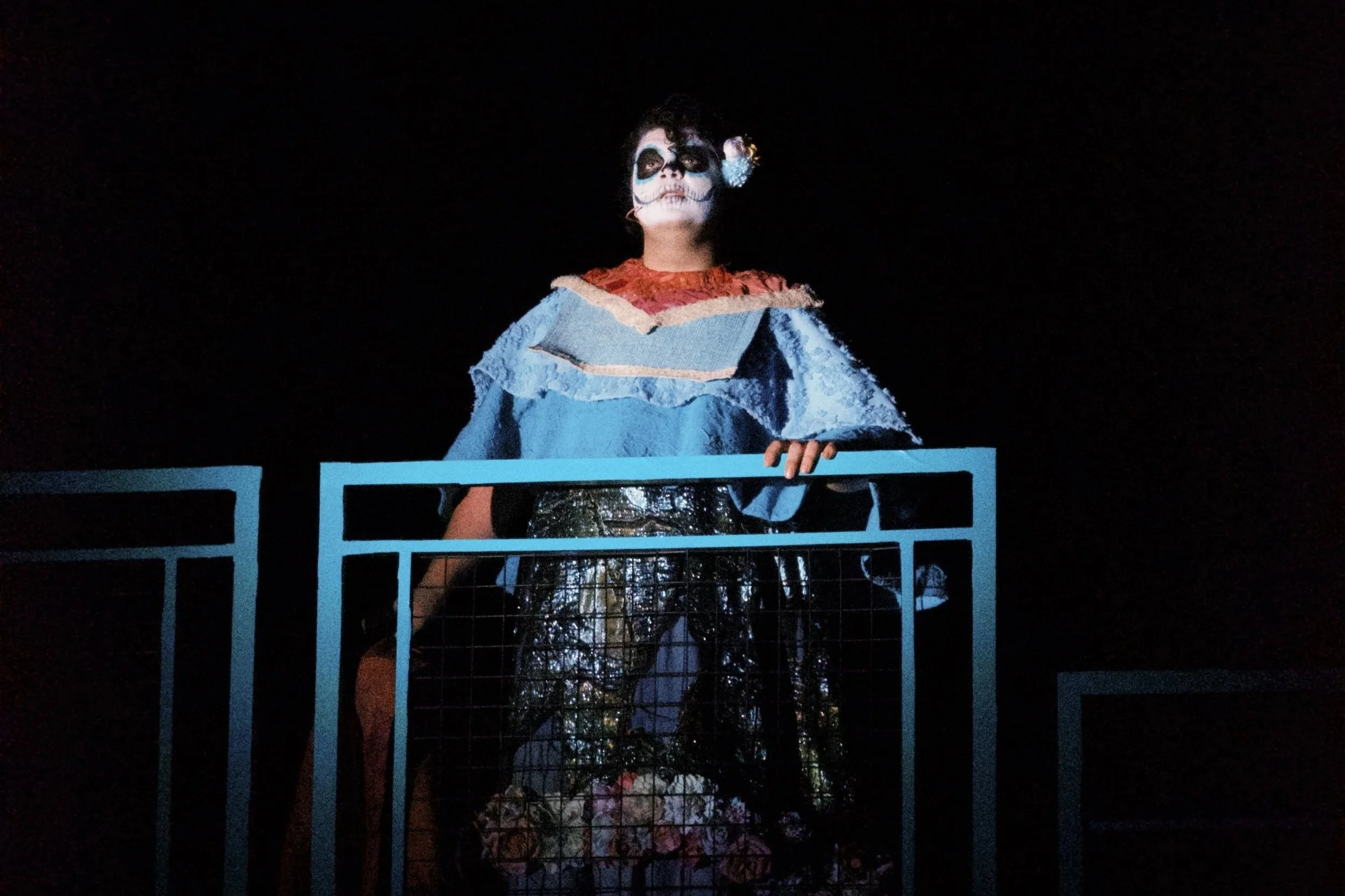 A person dressed in a colorful costume with face paint resembling a skeleton or Day of the Dead makeup, standing behind a blue metal railing at night.