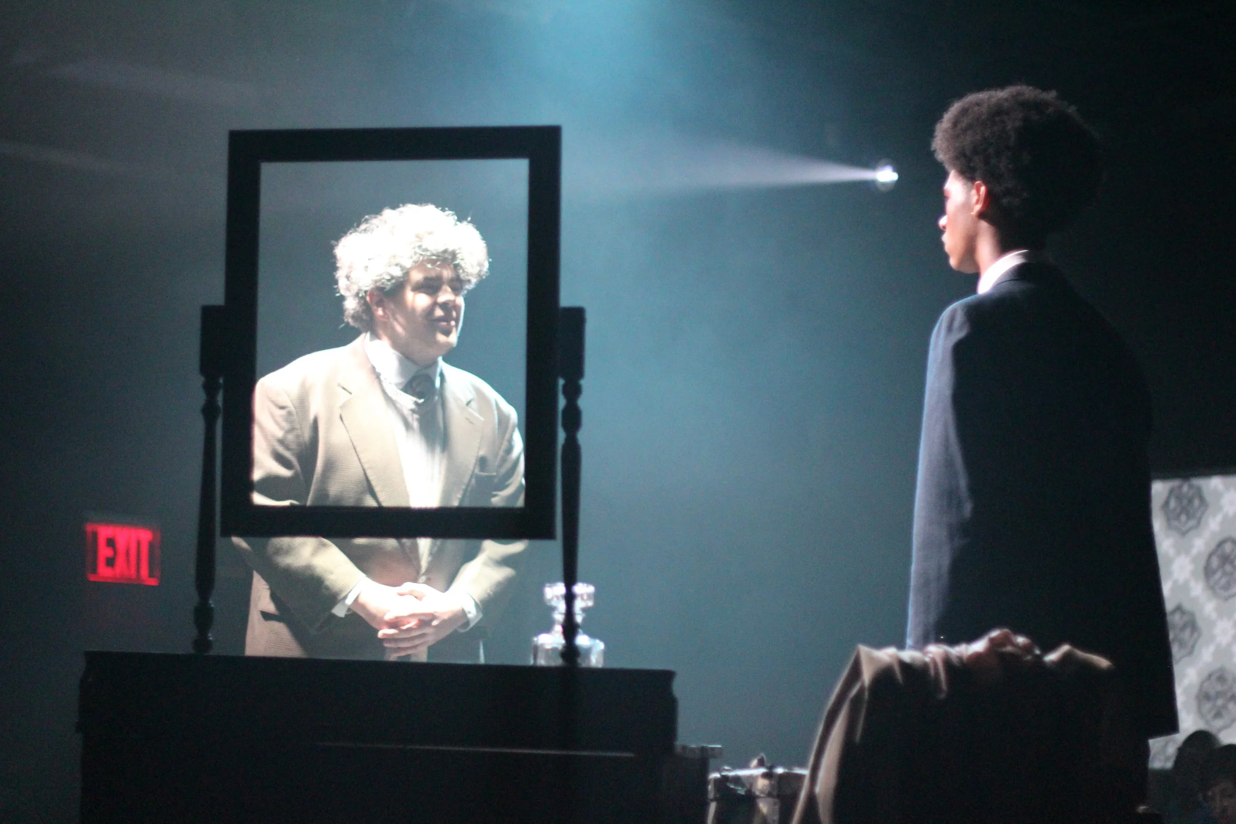 A young man with an afro hairstyle looking at his reflection in a mirror, while an older man with curly gray hair is seen in the mirror's reflection. They are in a dimly lit room.