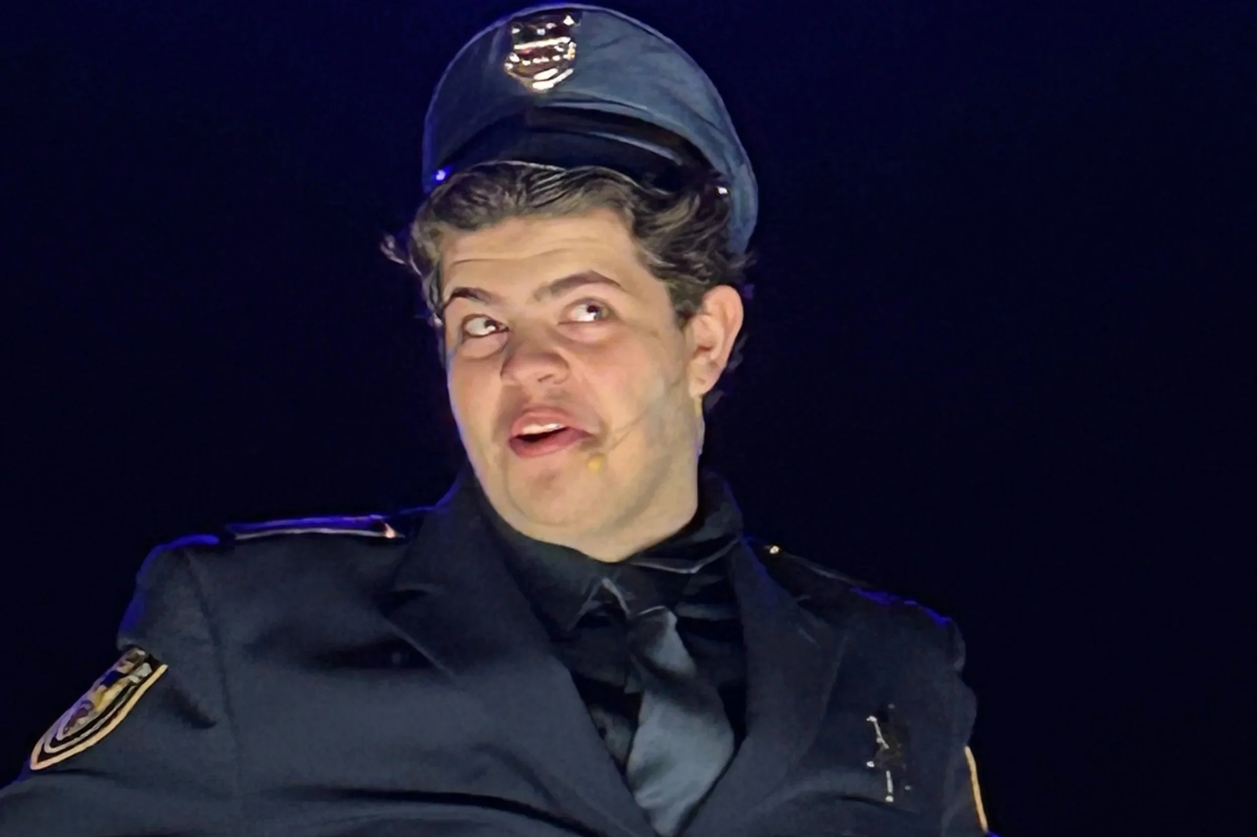 A man dressed as a police officer with a police hat, black uniform, and badge, making a funny facial expression with his eyes rolled and mouth slightly open against a dark background.