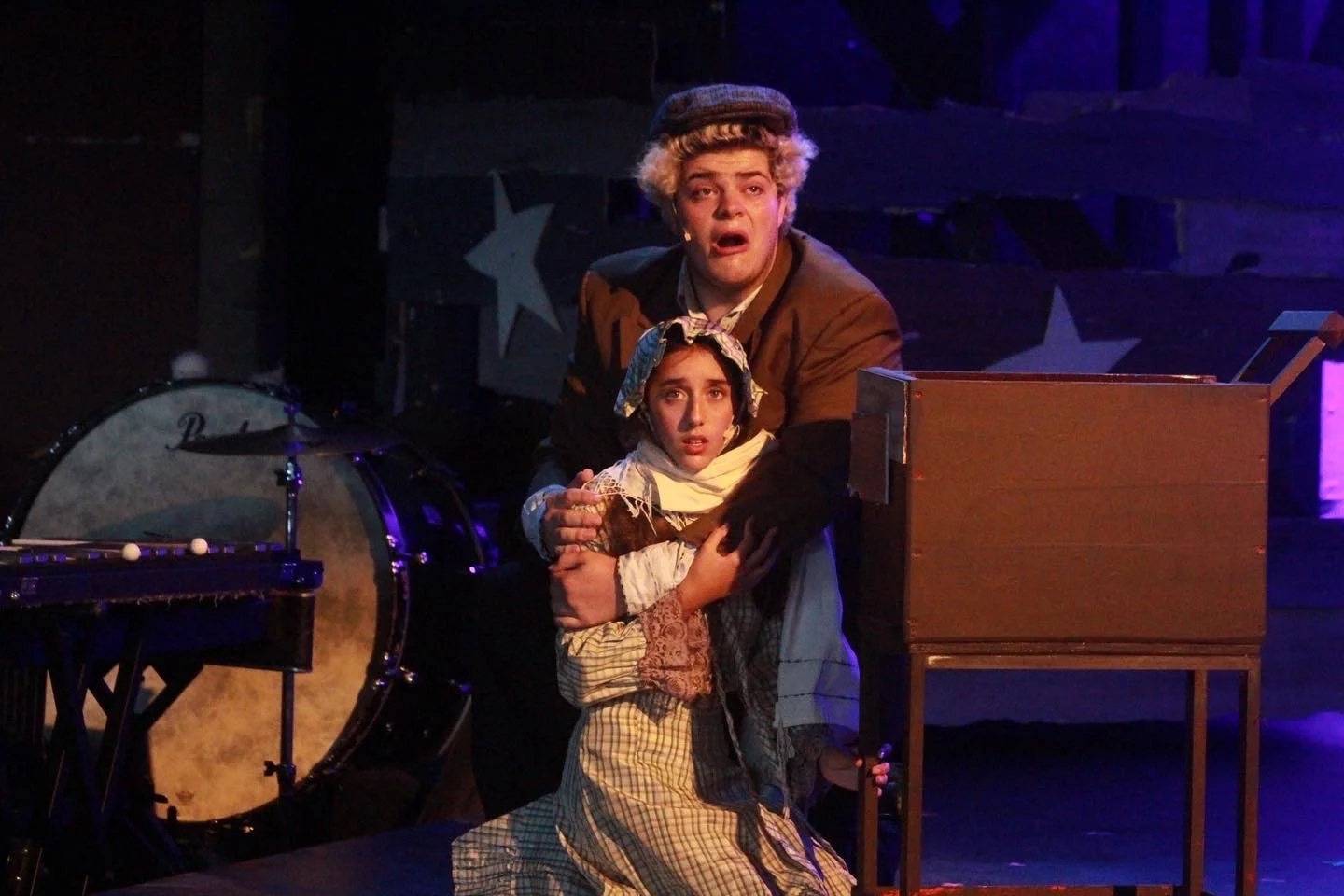 Two actors on stage, one adult male and one young girl, both with distressed expressions, dressed in vintage costumes, surrounded by stage props and musical instruments, suggesting a theatrical performance.