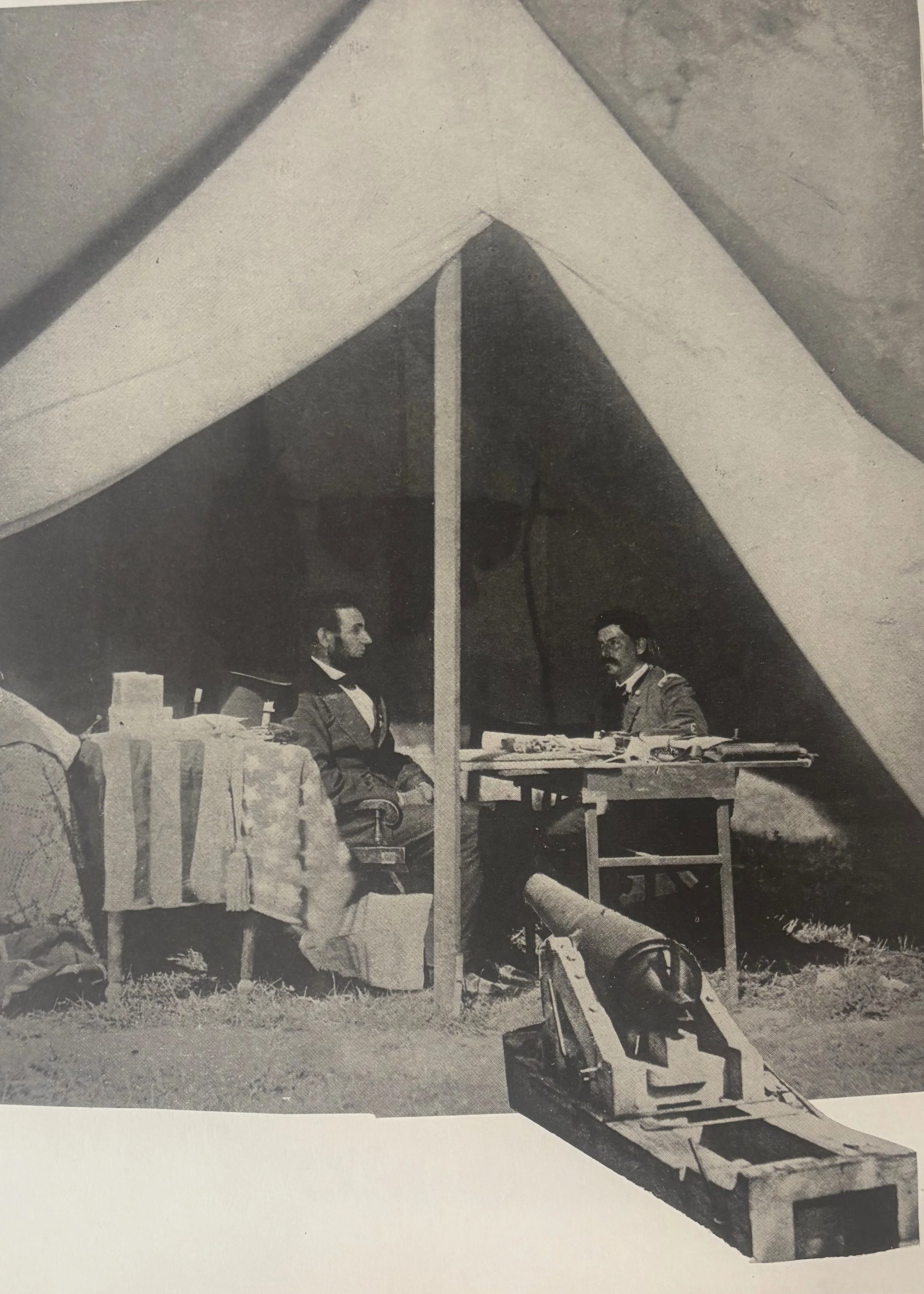 Historical black and white photo of two men in 19th-century military uniforms sitting inside a tent, with a table and a cannon in front.