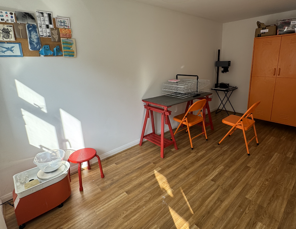 A room with a wooden floor, a bulletin board with crafts and photos, a small red stool, a table with adjustable red legs and a gray top with a wire mesh organizer, two orange folding chairs, a small black folding table with a machine on it, and an orange cabinet with storage boxes on top. Sunlight casts shadows on the wall.