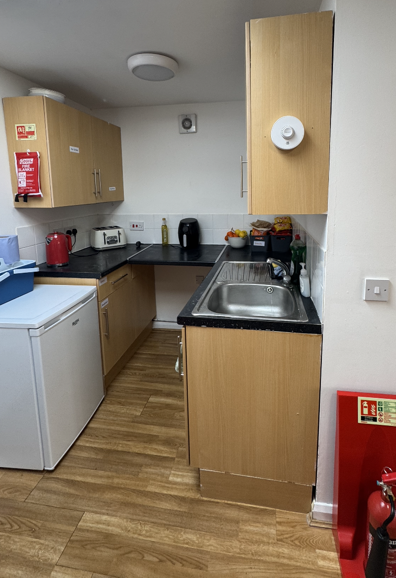 Small kitchen with wooden cabinets, a sink, countertop appliances including a red kettle, a toaster, and an air fryer, with fruit and kitchen supplies on the countertop. There is a white refrigerator and a fire safety box nearby.