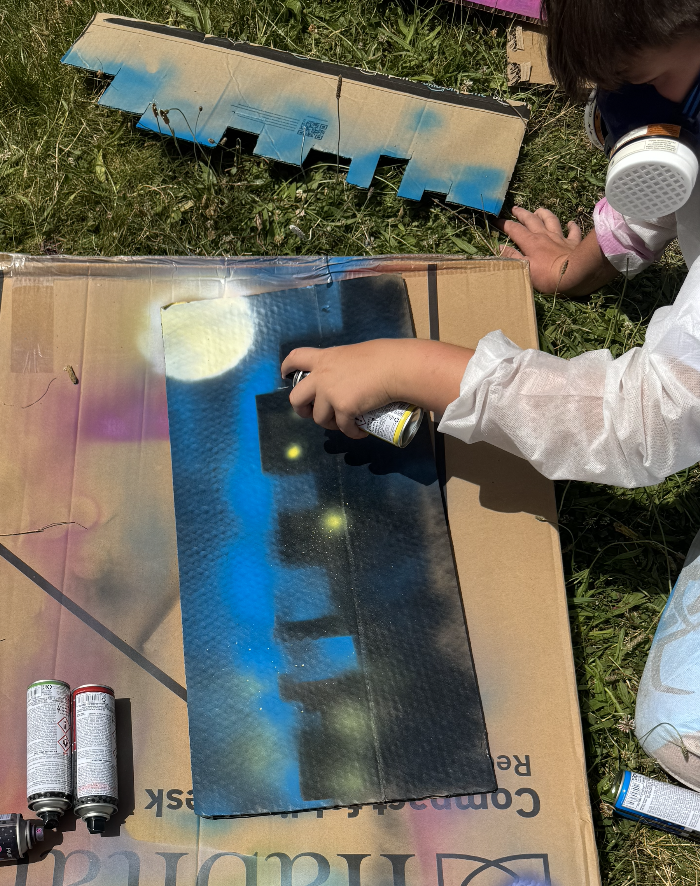A child wearing a white protective suit and a respirator mask is spray painting a panel sheet with spray paint cans outdoors on a grassy area. The panel has a painted sky scene with clouds and stars.