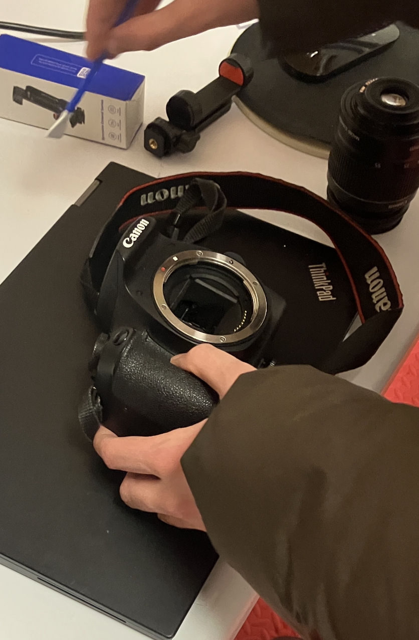 Person installing a camera lens into a Canon camera body on a table, with camera accessories and a box nearby.