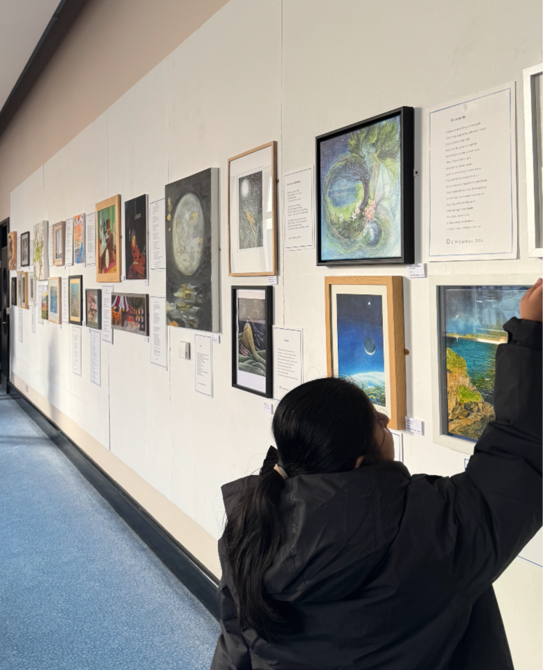Pupil with long dark hair in a ponytail wearing a black jacket, viewing artwork displayed on a gallery wall.