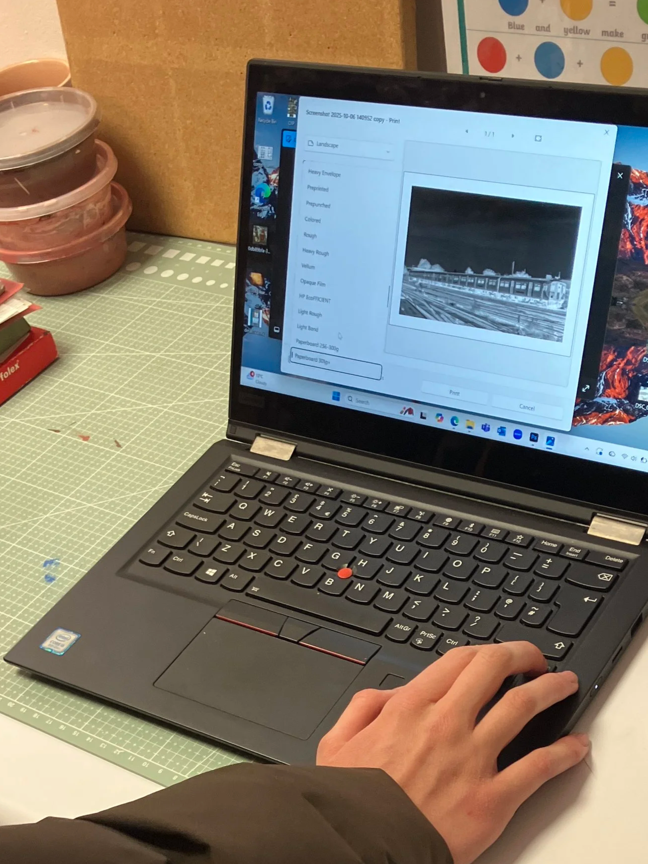 A person is working on a laptop at a work or craft table, with several small plastic containers stacked on the side, and a corkboard in the background. The laptop screen displays a printing dialog with a black and white photo of a train yard.