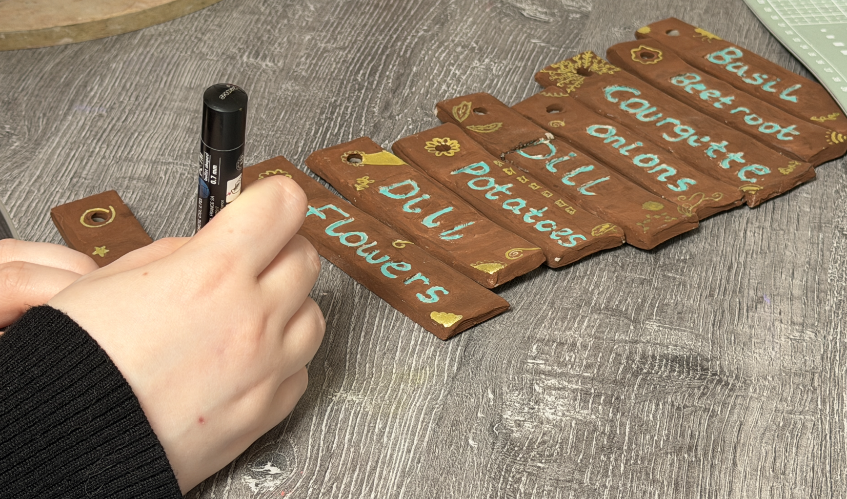 Hand holding a black marker, writing on small wooden signs painted with colorful letters and decorative gold patterns, with more signs laid out on a textured gray wooden surface.