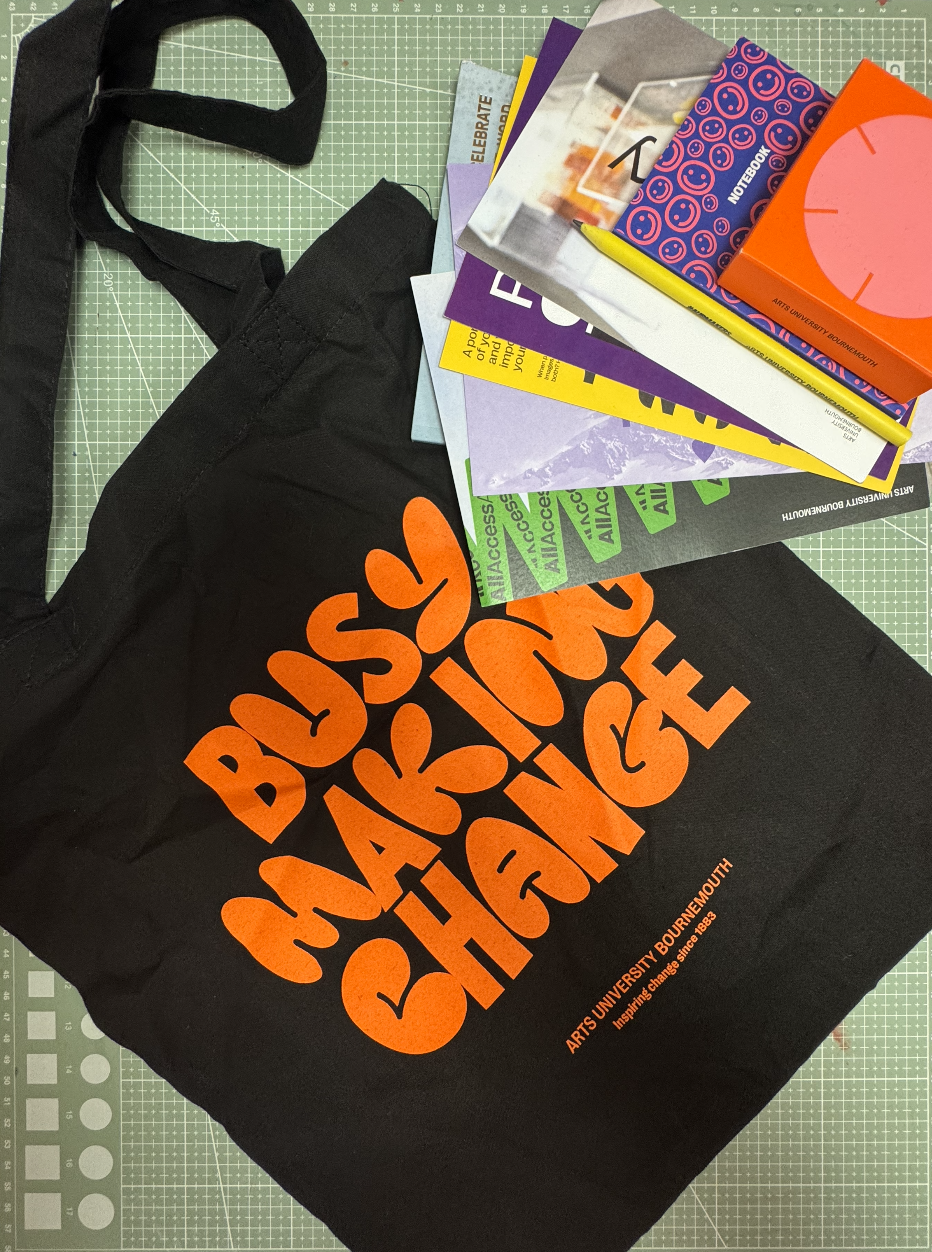 A black tote bag with orange text that reads 'ARTS UNIVERSITY BOURNEMOUTH' on a green cutting mat, surrounded by various colourful notebooks, a yellow pen, and paper.