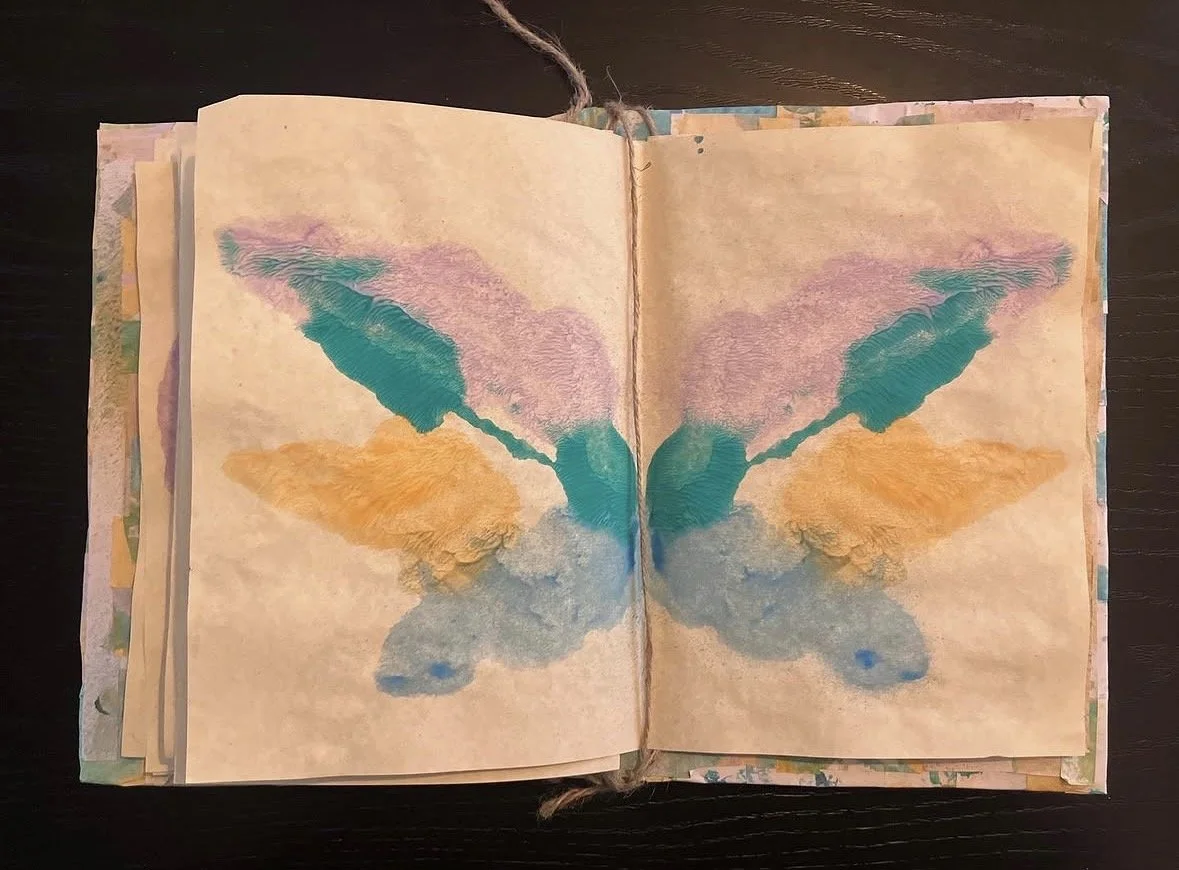 A watercolor painting of a butterfly with symmetrical wings in shades of teal, pink, orange, and blue on open pages of a sketchbook.