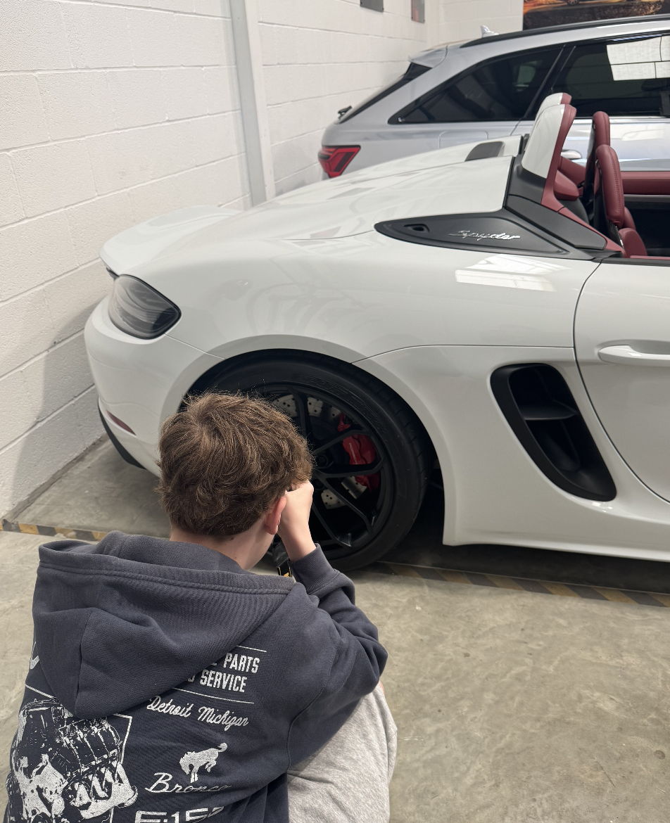 A pupil crouched on the floor taking photos of a white sports car with a black wheel and a red brake caliper, parked indoors near a concrete wall.