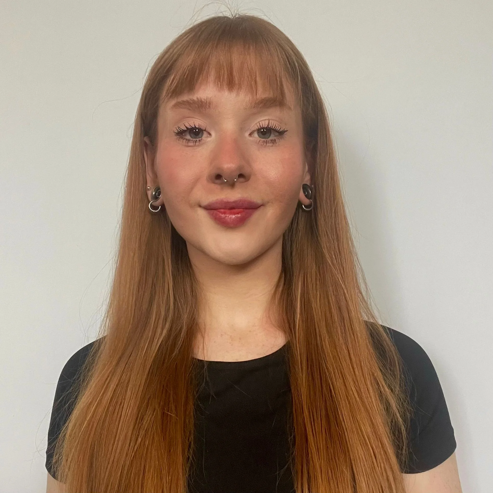 A young woman with long red hair, tattoos, multiple earrings, wearing makeup, smiling slightly, against a plain light background.