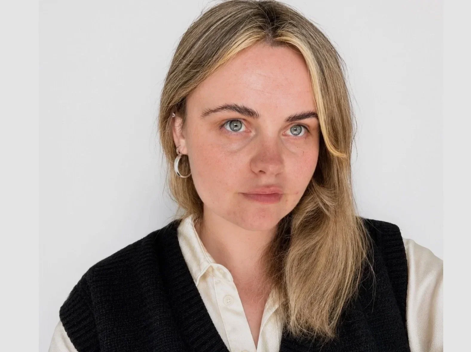 Close-up photo of a woman with blonde hair, blue eyes, wearing silver hoop earrings, a white collared shirt, and a black sweater vest, against a plain white background.