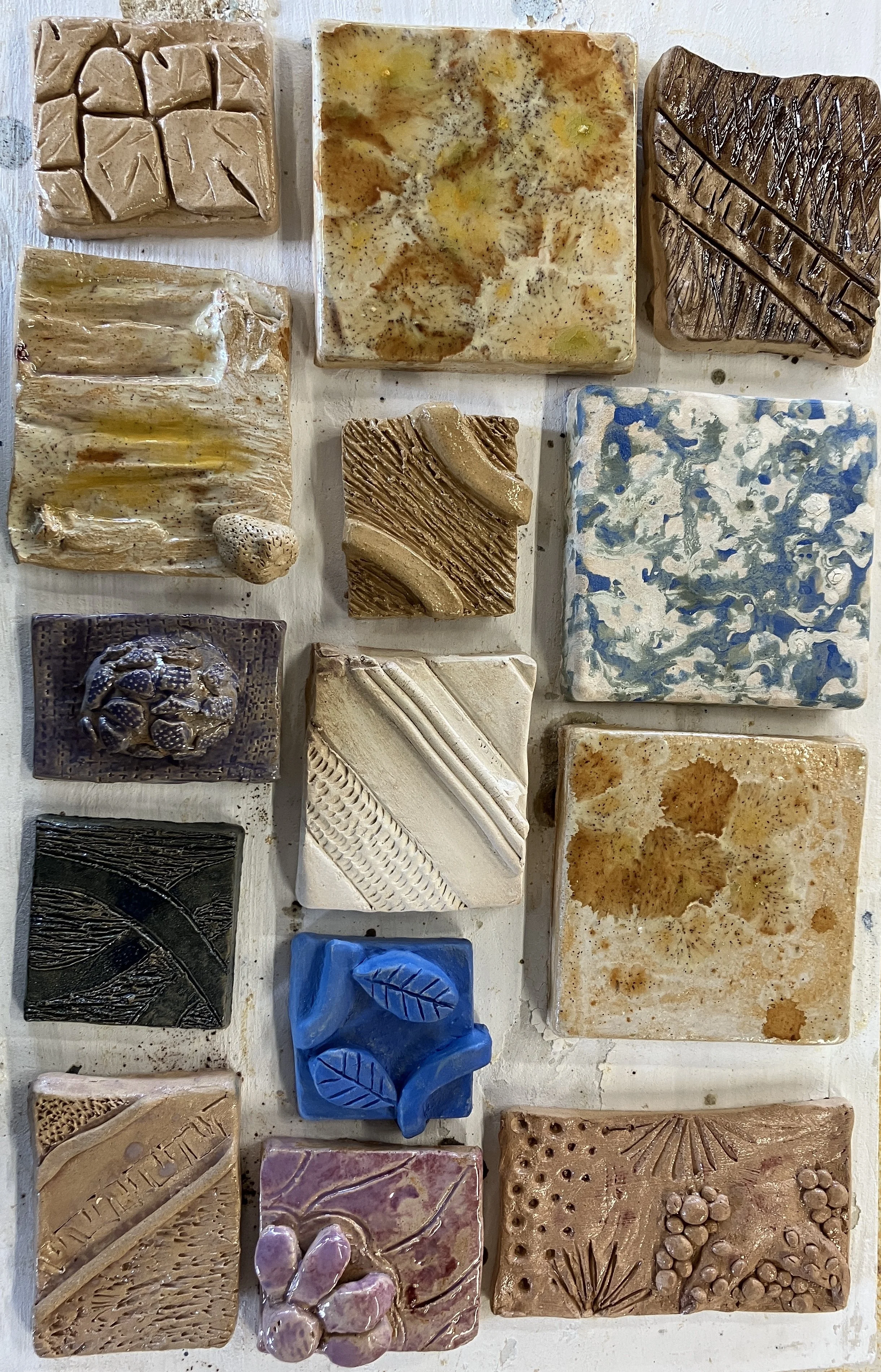Collection of various handcrafted tiles with different colors, textures, and decorative patterns arranged on a white surface.