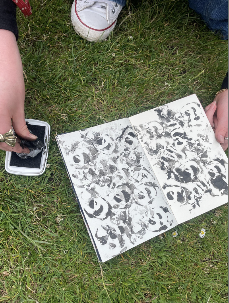 Person creating a printmaking artwork on grass, using a roller and black ink, with a print of abstract face images on paper.