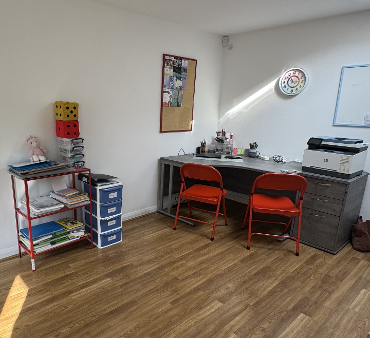 A small office or classroom with a wooden floor, white walls, and a window letting in sunlight. There are two red chairs at a desk with various office supplies, a printer on a gray cabinet, a bulletin board on the wall, and a red shelving unit with binders, children's books, and plush toys.