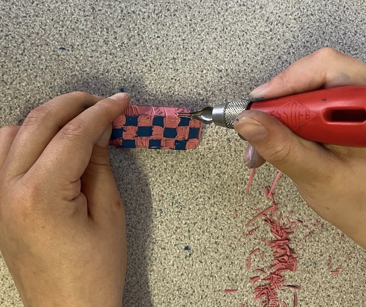 Hands creating a woven pattern on a small rectangular object using a rotary tool, with pink and blue strips and wood shavings on a beige surface.