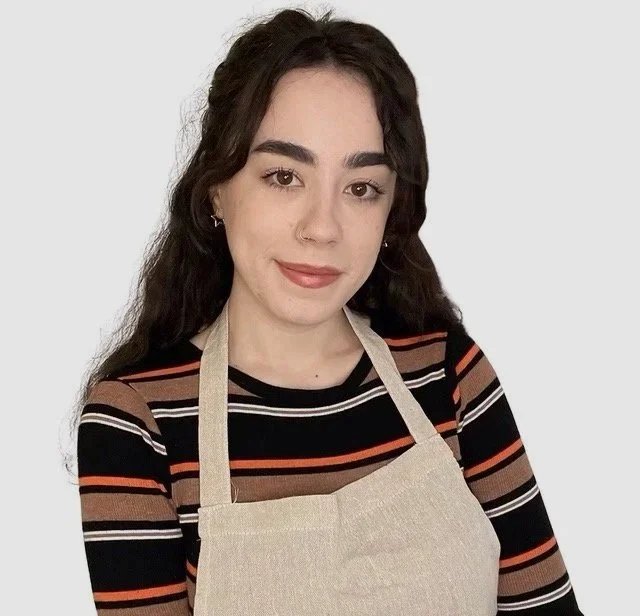 Young woman with long, wavy dark hair wearing a striped long-sleeve shirt and a beige apron, smiling. Plain white background.