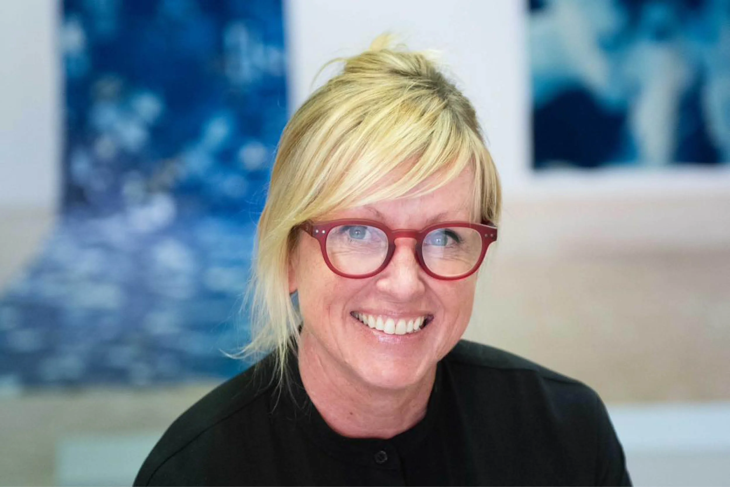 Close-up of a smiling woman with short blonde hair and red glasses, indoors with blue abstract artwork in the background.