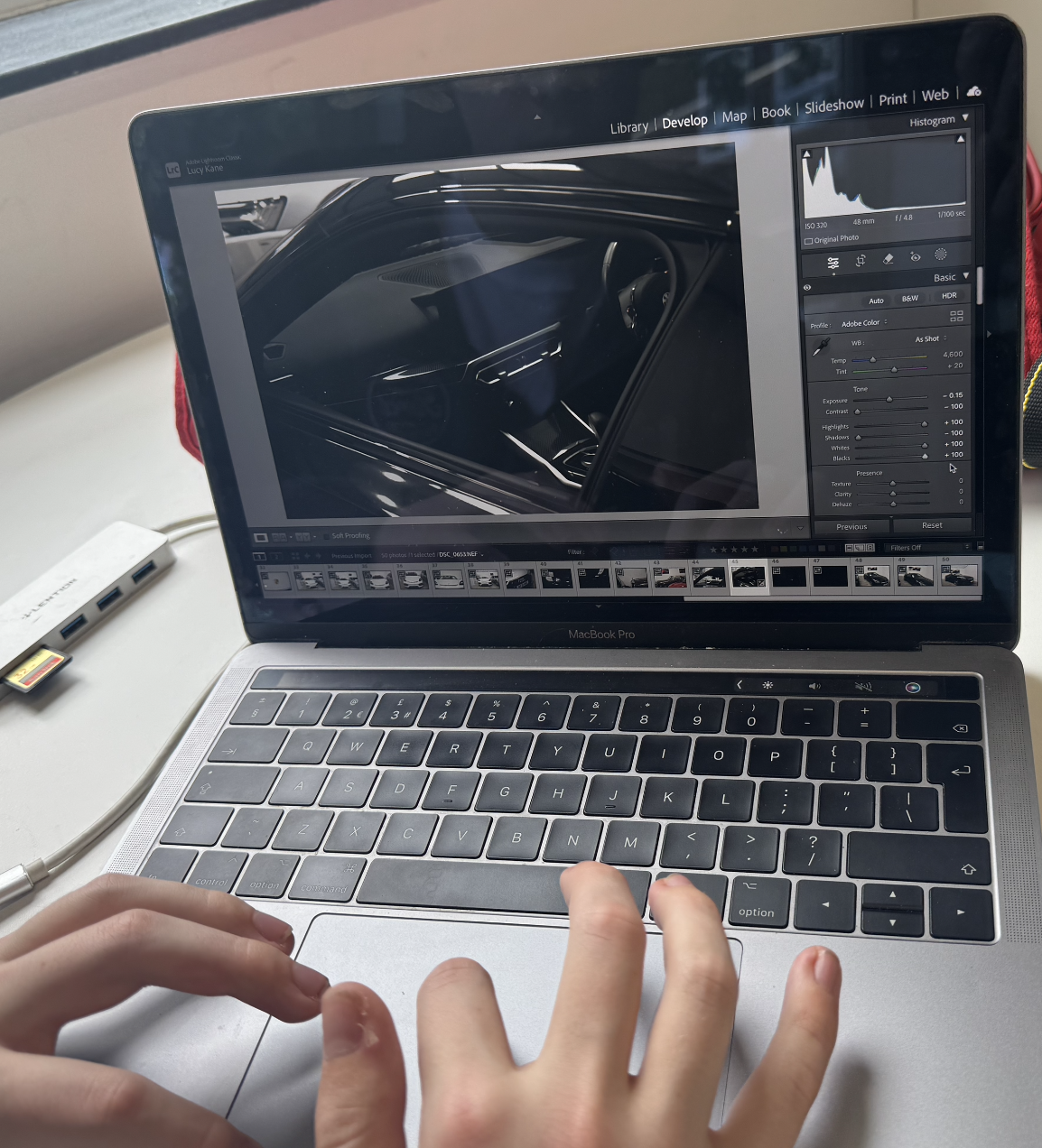 A person editing a photo of a car's interior on a MacBook Pro laptop using photo editing software, with a docked USB-C hub nearby.