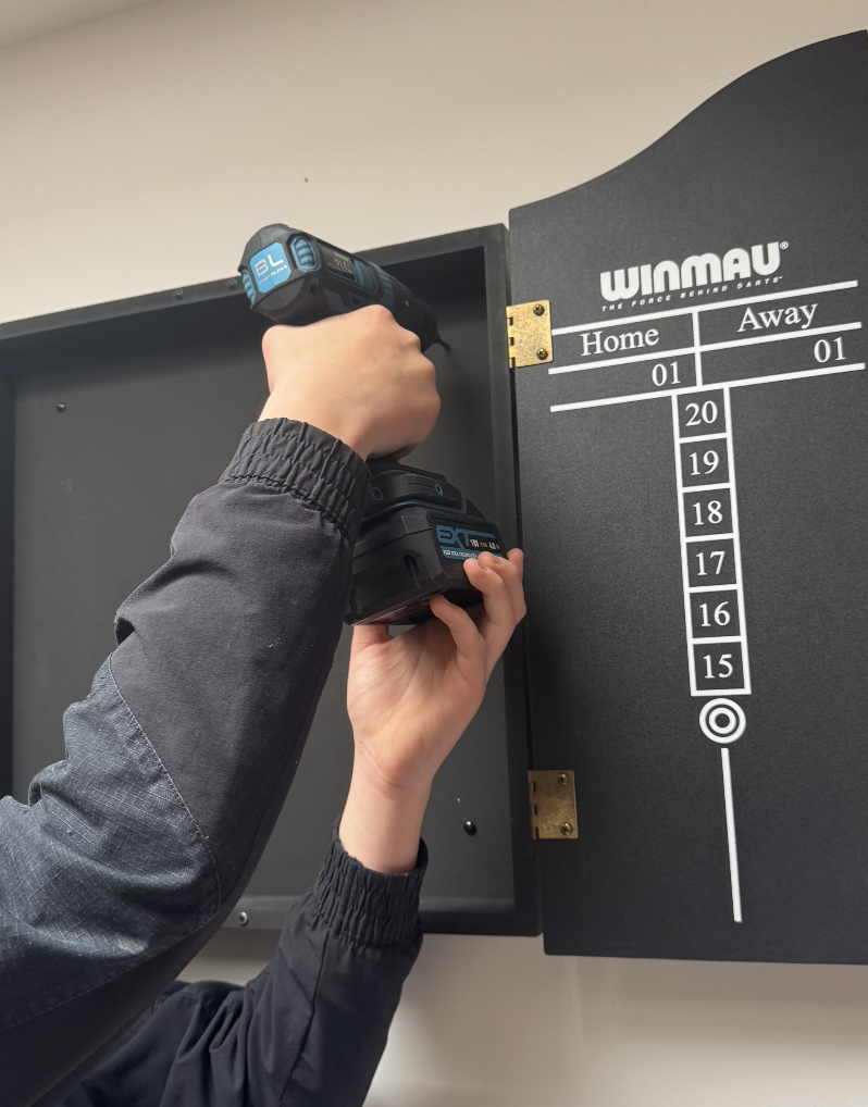 Person using a cordless drill to attach a l black cabinet with a scoreboard on top, for a darts game.