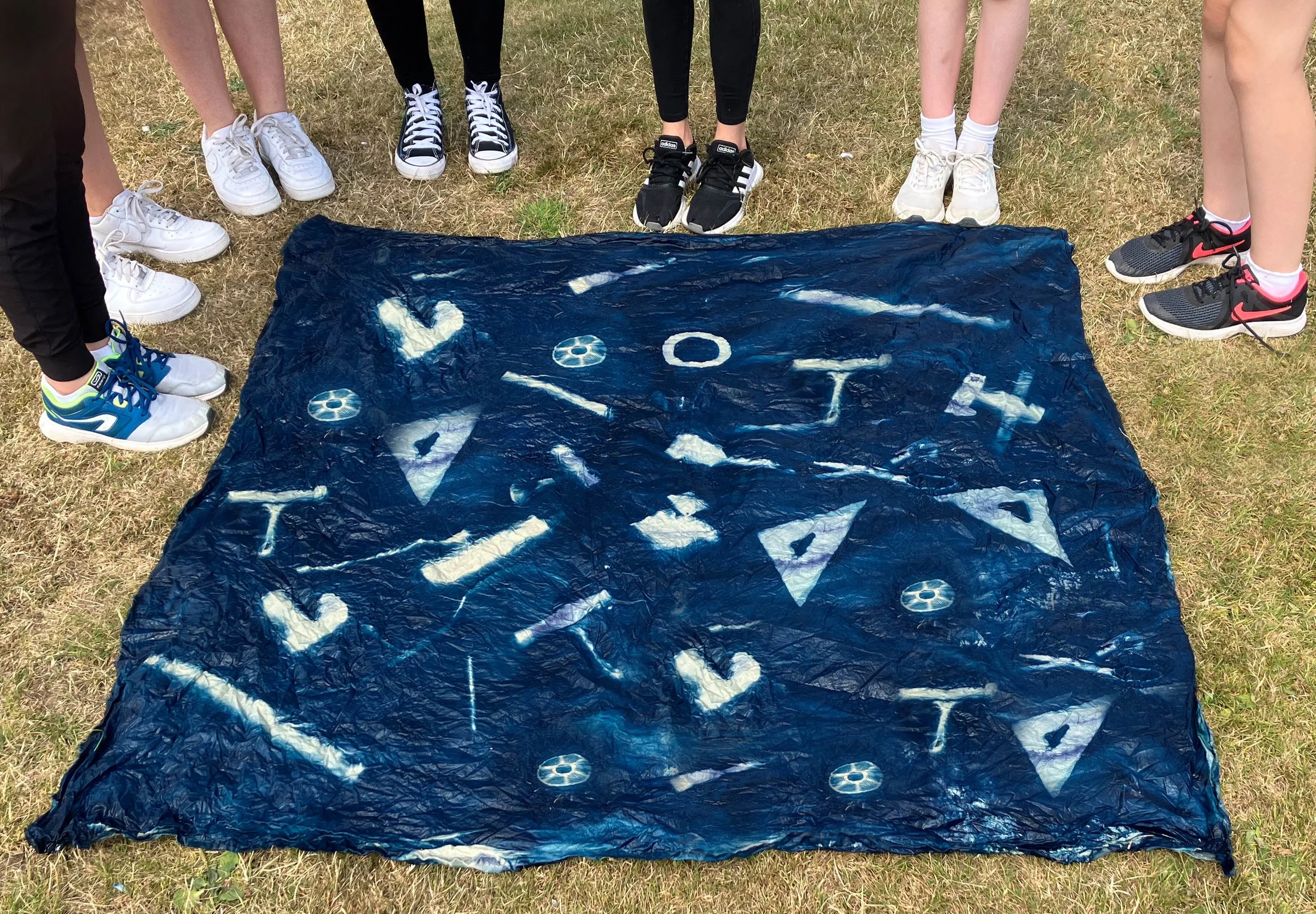 People standing outdoors on grass in a circle, wearing sneakers, surrounding a blue tarp with white markings on the ground.