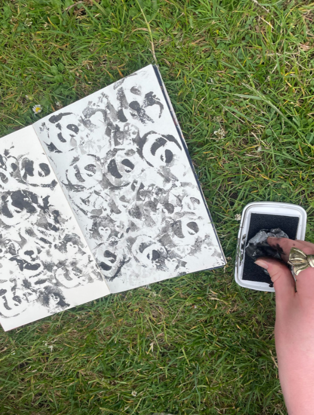 Open sketchbook with multiple black ink stamps on pages, placed on grass, with a hand holding a rubber stamp and ink pad nearby.