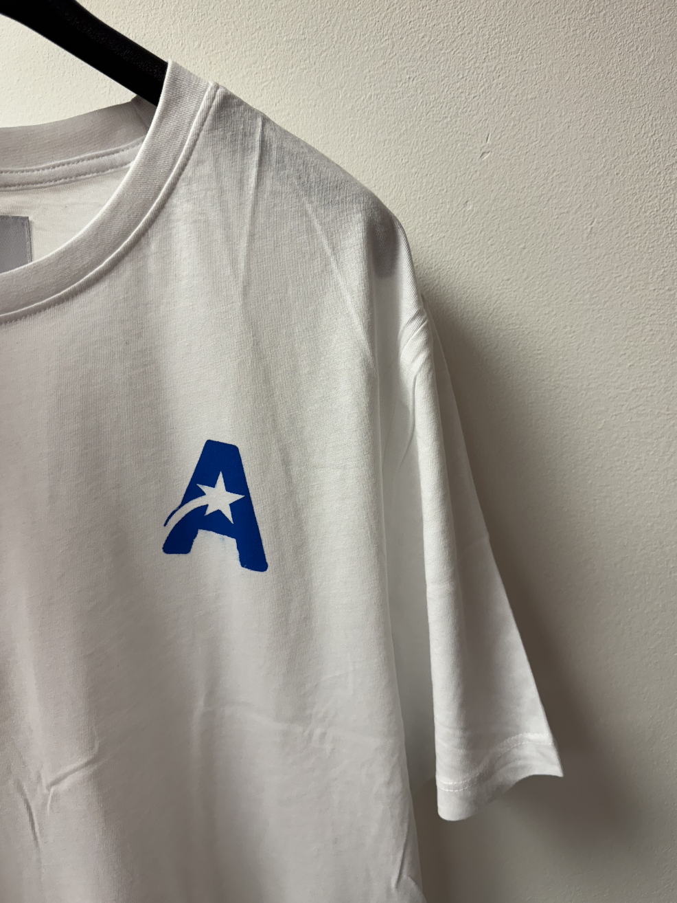 White T-shirt with a blue logo featuring a star and a stylized 'A' hanging on a black hanger against a beige wall.