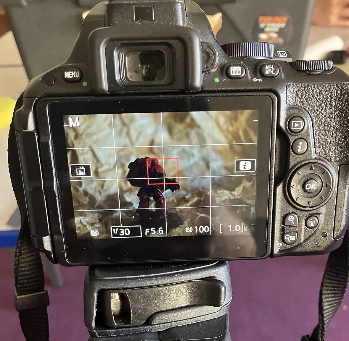 A DSLR camera on a tripod capturing a close-up photo of a small figurine with a backpack, set against a textured earthy surface.