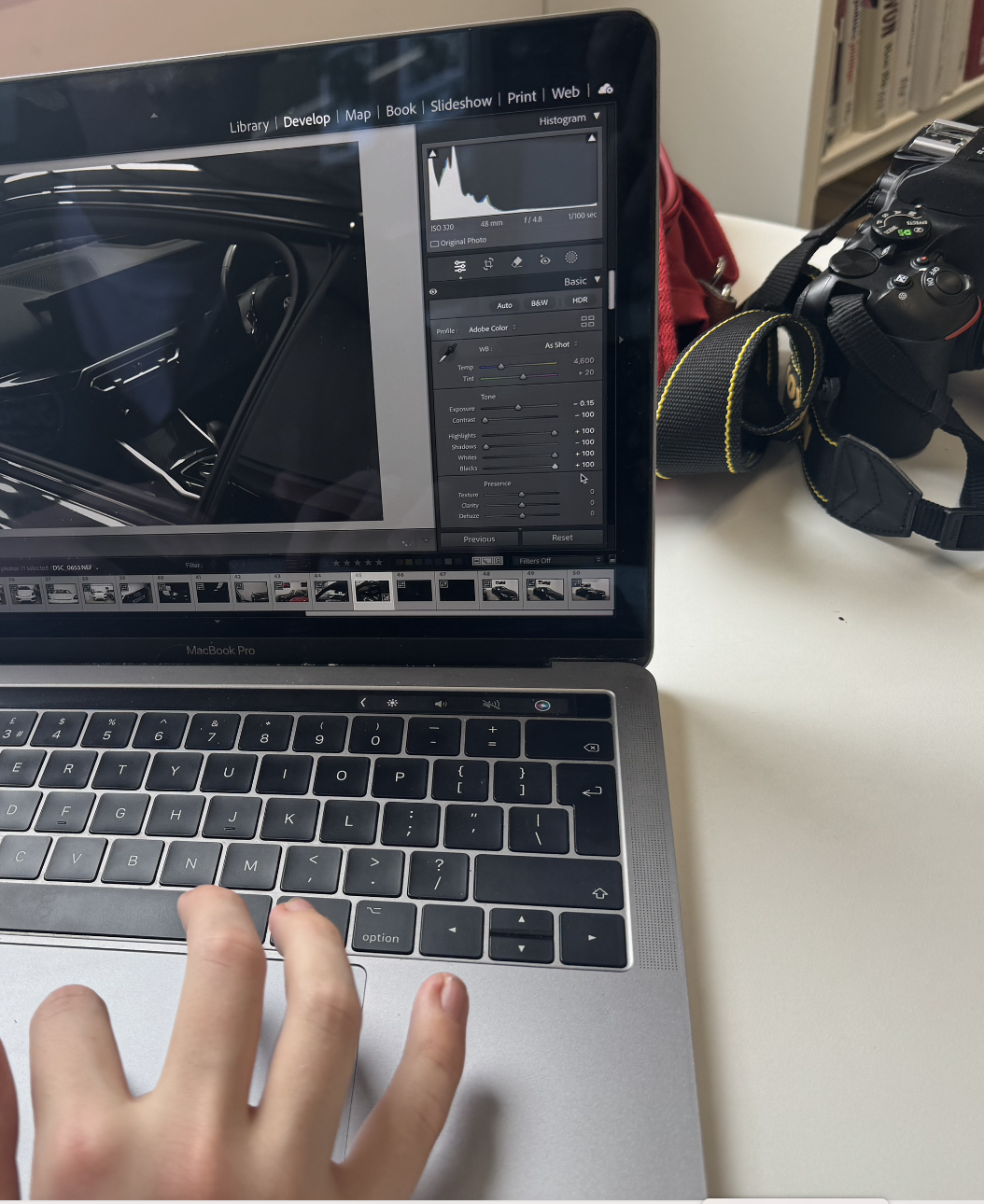 A laptop on a desk displaying photo editing software with histogram and adjustment sliders, a hand with three fingers is touching the trackpad, and a DSLR camera with a strap is beside the laptop.