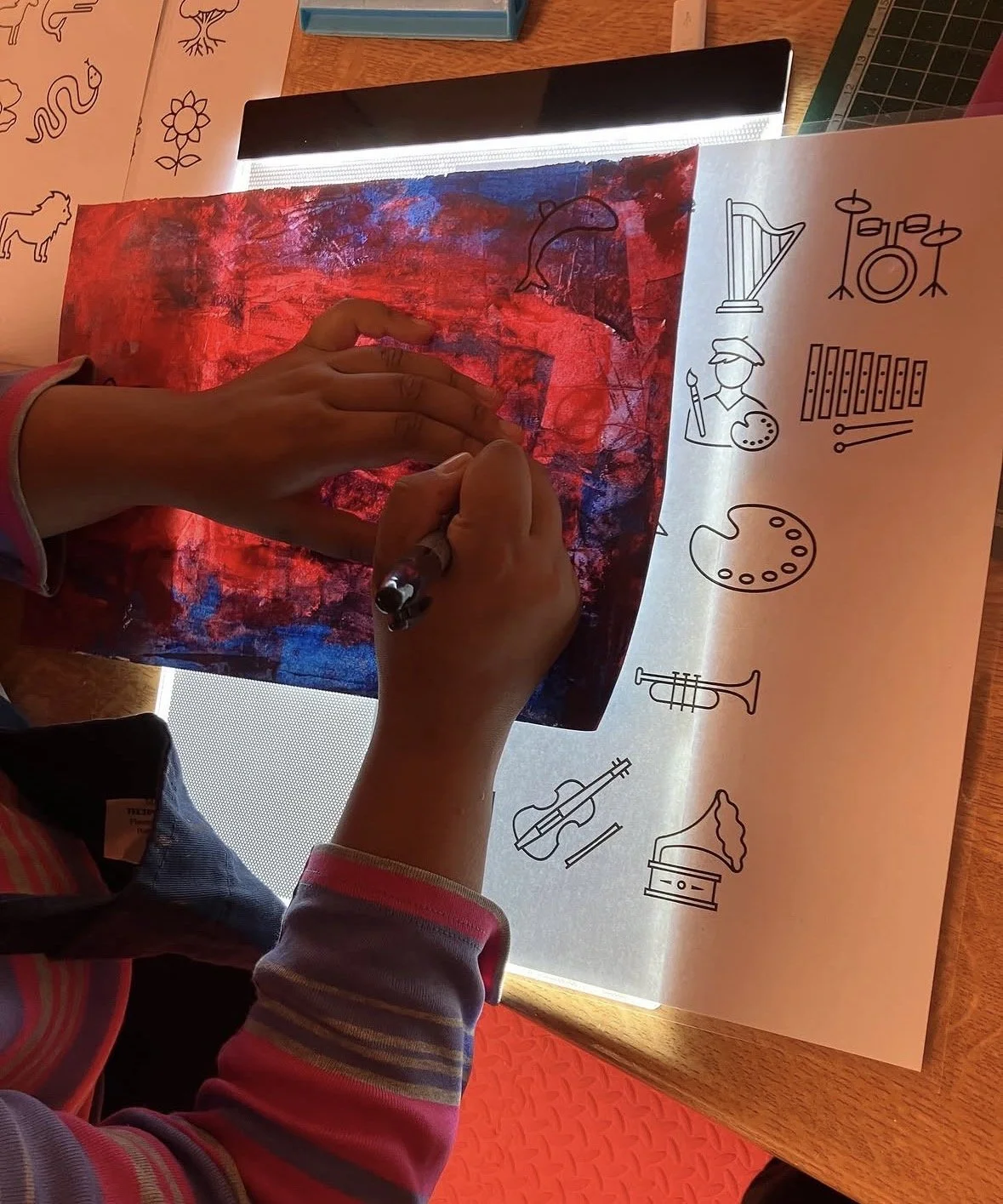 A person is creating a mixed media art piece by placing a transparent sheet with stenciled black line drawings of musical instruments, a paintbrush, and a person painting, over a colorful background with red, blue, and purple hues. The person's hands are visible, one holding a pen and the other pressing the sheet onto the artwork. The background is illuminated from behind, highlighting the textured abstract art underneath.