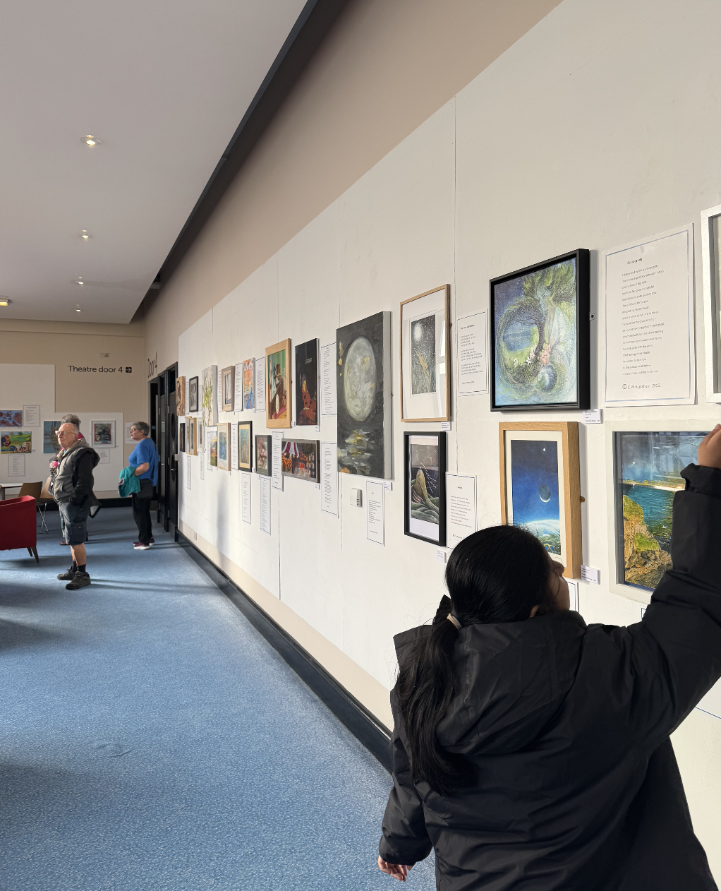 People viewing artwork on a gallery wall inside an art exhibit, with paintings and descriptions displayed.