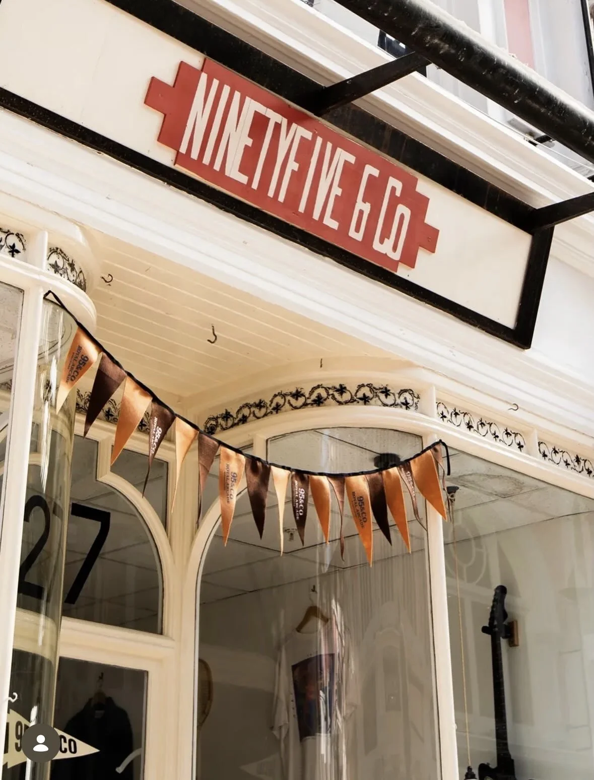 Street sign reading 'NINET YFIVE 60' mounted above a storefront with a decorative black iron railing, orange and black Halloween-themed pennant banner hanging in front, window with white decorative trim, and various items partially visible.