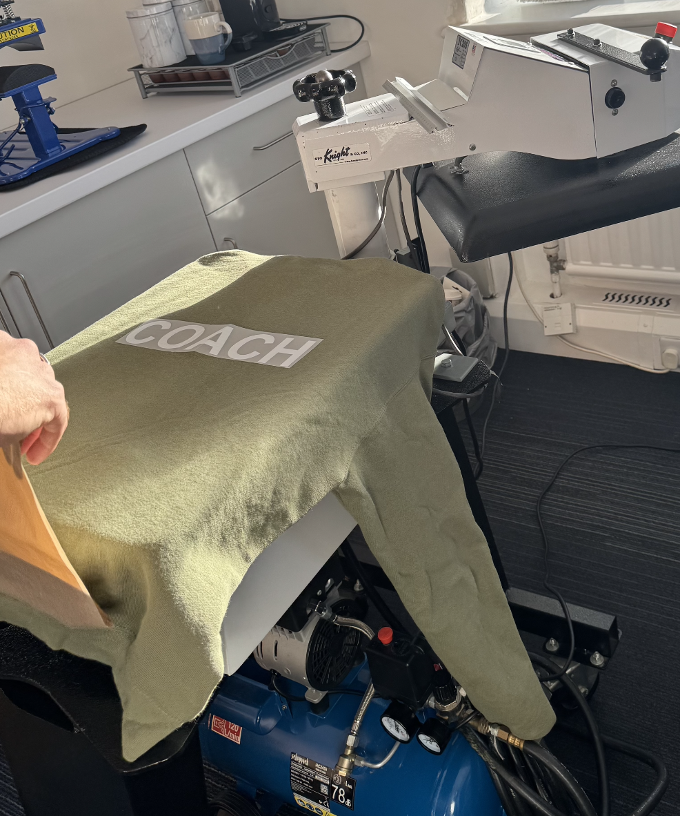 T-shirt Printing machine, heat press, and a piece of fabric with the word 'COACH' on it in a tattoo studio or nearby workspace.