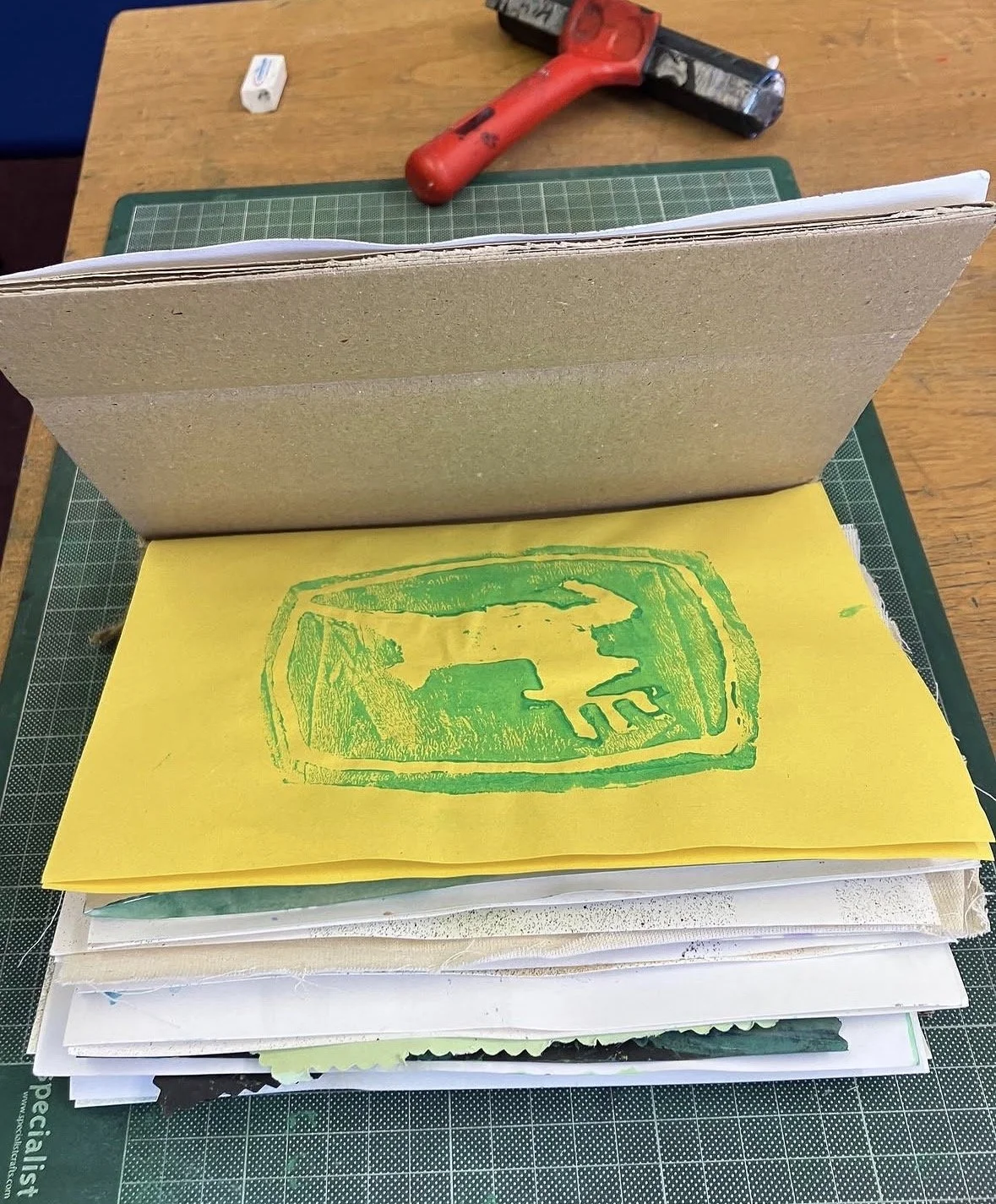A work desk with a pile of papers and a yellow paper with a green print of a Deer silhouette inside a rounded rectangle. There is a red roller, a white eraser, and a green cutting mat on the desk.