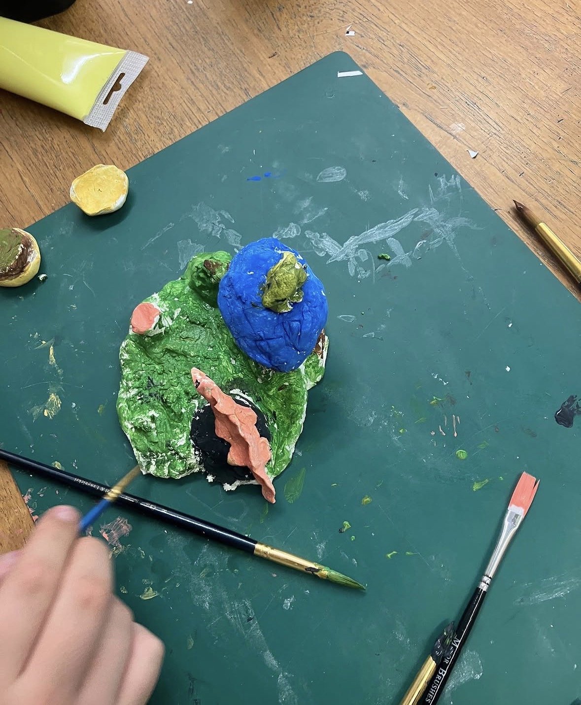 Colorful clay sculpture of a green crocodile with a blue head and pink tongue on a green work surface, surrounded by art supplies.