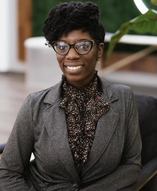 A woman with short curly hair, glasses, and a smile, wearing a gray blazer and patterned blouse, sitting indoors with a plant and modern decor in the background.