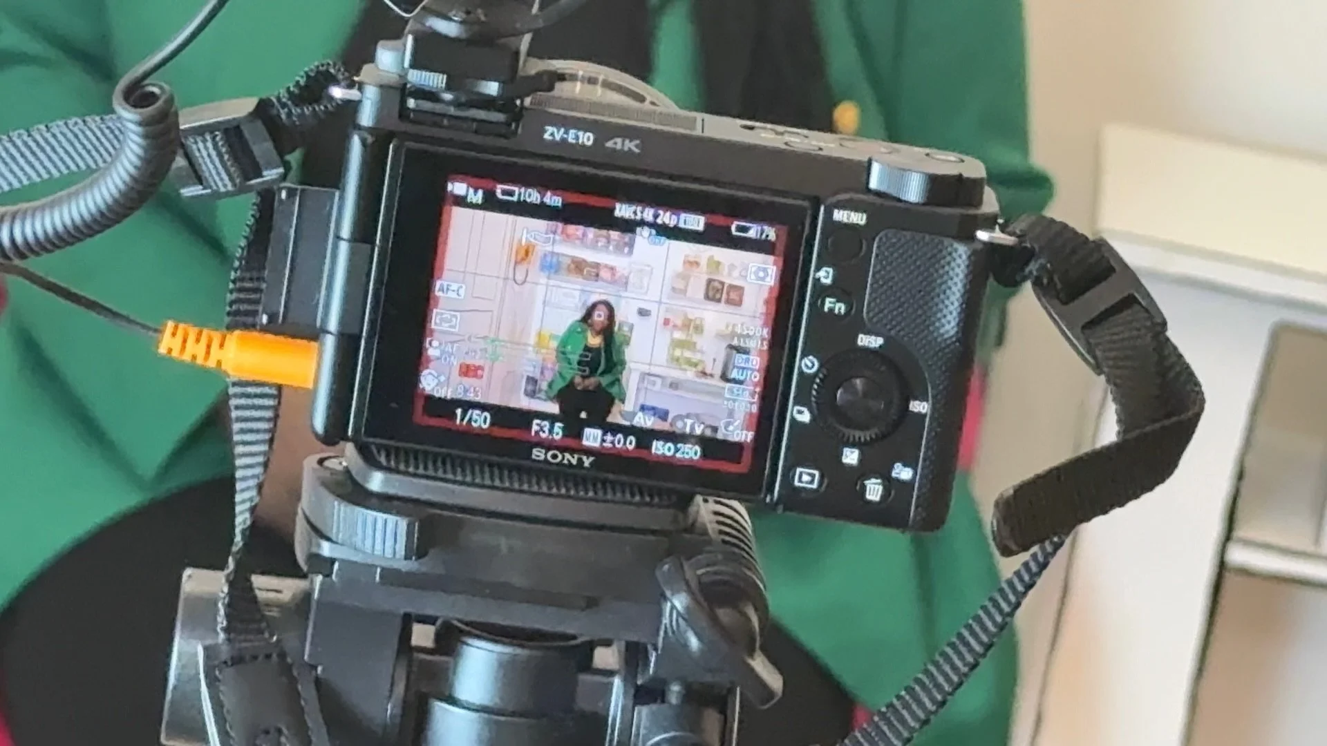 A camera mounted on a tripod captures a woman standing in front of a colorful Background of shelves and drawers.