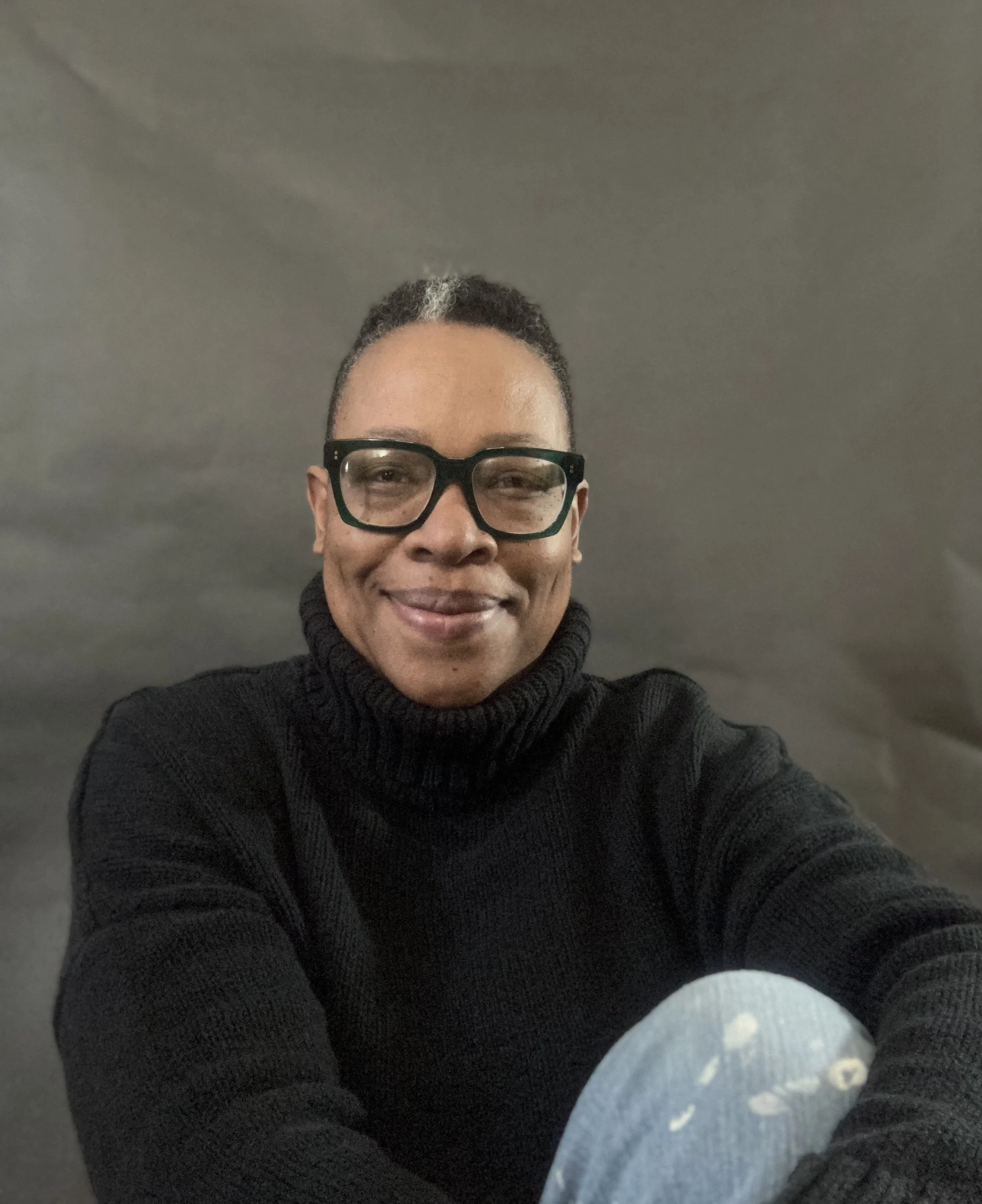 A person with short hair, wearing glasses and a black turtleneck sweater, smiling at the camera.